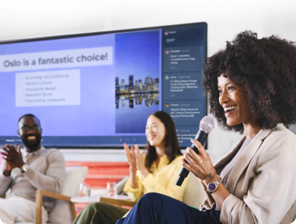 A woman with curly hair smiles while holding a microphone at a team event. There is a large presentation screen behind her with 2 colleagues sat in front of it.